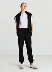 Pantaloni da jogging neri con polsini elasticizzati, abbinati a una maglietta bianca. Un maglione nero è drappeggiato sulle spalle. Scarpe chiare completano il look.