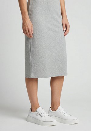 Person iført et knælængt lysegråt skørt og hvide lave sneakers, stående mod en ensartet lys baggrund.