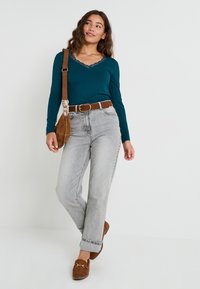 Top de manga larga color verde azulado con cuello de encaje, jeans de cintura alta grises, cinturón marrón y mocasines de ante marrones; llevando un bolso crossbody marrón.