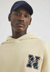 Kremowa bluza z kapturem z haftowanym granatowym "M" i logo. Noszona z ciemnoniebieską czapką z korka. Gładka tekstura materiału, minimalistyczny design.