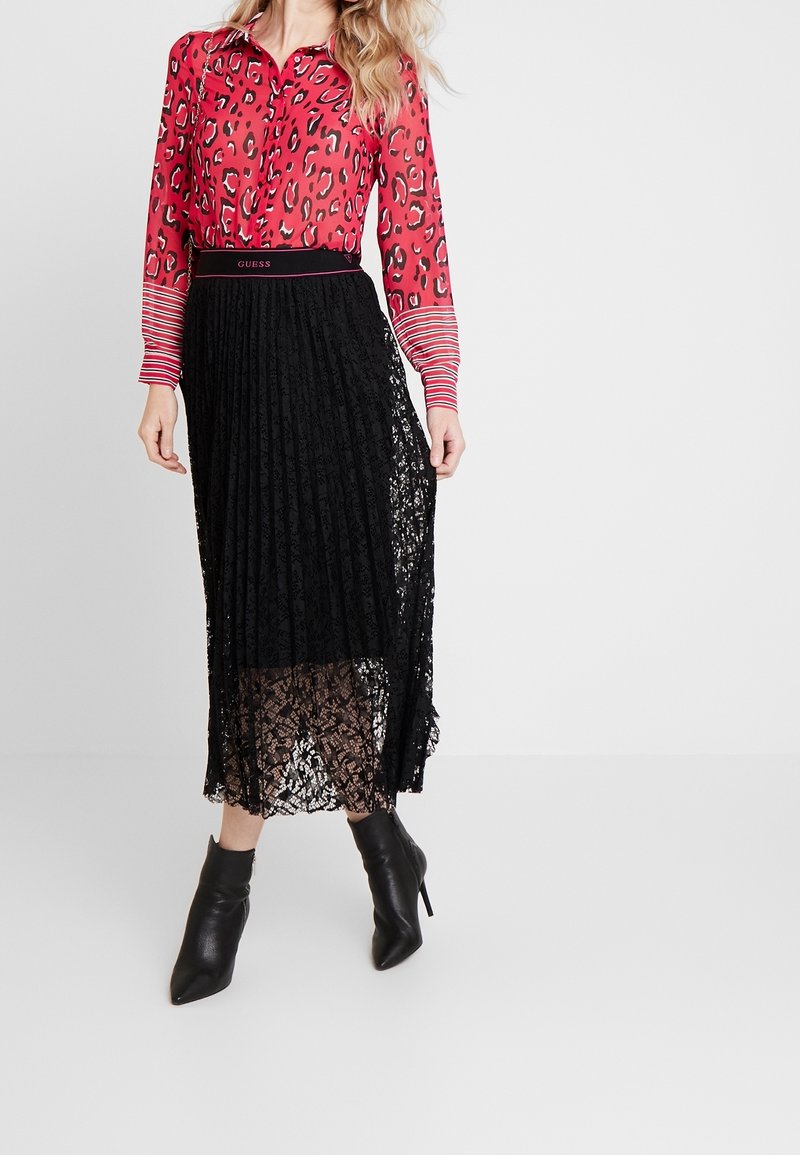 Femme portant un chemisier rouge à motif léopard, une jupe midi plissée en dentelle noire avec une ceinture Guess, et des bottines à talons noires, debout devant un fond blanc.