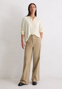 Lichtgeel button-up shirt met lange mouwen, gecombineerd met beige wijde broek en zwarte laarzen. De stof lijkt glad en zacht.