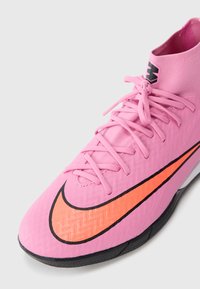 Scarpa da calcio alta rosa con logo Nike arancione brillante, lacci rosa e suola testurizzata su sfondo bianco.