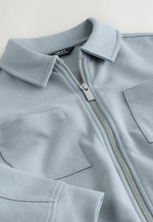 Svetlomodrá bunda na zips vyrobená z mäkkej látky, s golierom, dvoma vreckami na hrudi a kovovým zipsom na zapínanie.