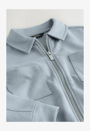 Giacca celeste con zip in tessuto morbido, dotata di colletto, due tasche sul petto e chiusura con cerniera in metallo.