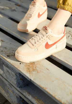 Stopy w beżowych i koralowych sneakersach Nike Air Force 1 spoczywają na postarzałej drewnianej palecie w świetle słonecznym.