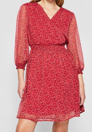 Femme portant une robe rouge à fleurs avec un col en V, des manches longues transparentes et une taille élastiquée froncée.
