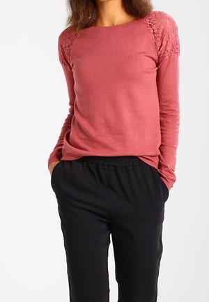 Frau trägt ein staubrosafarbenes Langarmshirt mit Spitzen-Details an den Schultern und schwarze Hosen mit elastischem Bund, Hände in den Taschen.