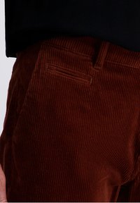 Brown corduroy pants with vertical ribbed texture. Features a side pocket and belt loops. The fabric appears soft and durable.