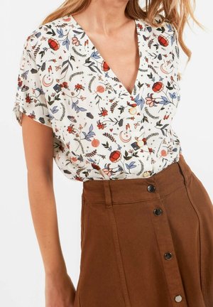 Vrouw draagt een witte blouse met korte mouwen met kleurrijke bloemen- en maanpatronen, gecombineerd met een hoog getailleerde bruine rok met knopen.