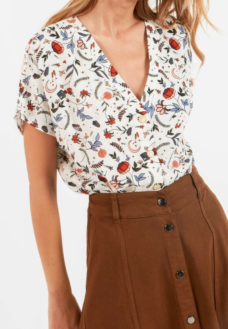 Femme portant un chemisier blanc à manches courtes avec des motifs floraux et lunaires colorés, assorti à une jupe marron taille haute boutonnée.