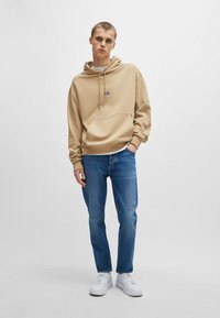 Beige hooded sweatshirt with a front pocket, paired with blue jeans and white sneakers, displayed on a model standing against a neutral background.