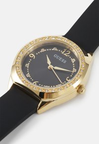 Gold-tone watch with a round case, black dial, crystal embellishments around the bezel, and a black silicone strap. Features Arabic numerals.