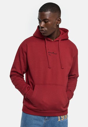 Homme portant un sweat à capuche rouge avec poche avant et un jean bleu, regardant vers sa gauche sur un fond uni.