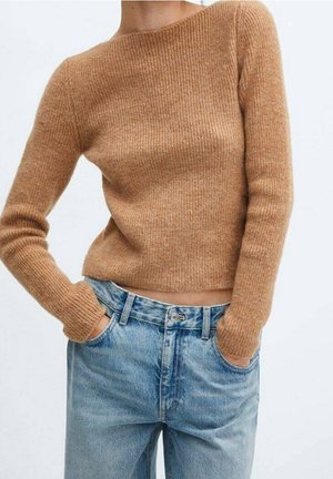 Person iført en tætsiddende, ribstrikket, kamelfarvet sweater og højtaljede lyseblå denimjeans med hænderne i lommerne.