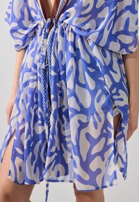 Vestido ligero y transparente con un patrón abstracto en azul y blanco, ajustado en la cintura con un lazo marrón y con aberturas laterales para facilitar el movimiento.