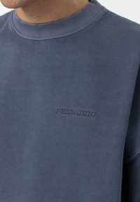 Mørkeblå sweatshirt med ribbet krave og broderet "PEGA-DOR" logo i matchende farve, med en blød, tekstureret stoffinish.