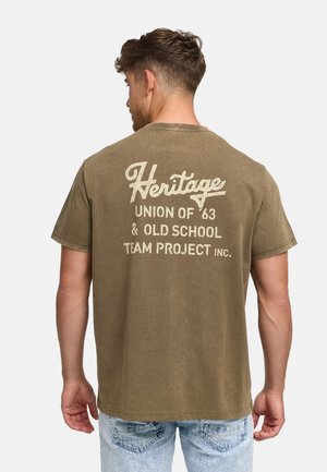 Man wearing olive green T-shirt with vintage white text "Heritage Union of '63 & Old School Team Project Inc." on the back, paired with light jeans.