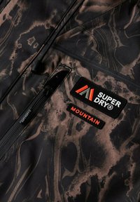 Close-up van donkergepatroneerde stof met een ritszak en twee patches met de teksten "SUPER DRY" en "MOUNTAIN" in vette letters.