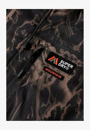 Zbliżenie na ciemną, wzorzystą tkaninę z zamkiem błyskawicznym i dwoma naszywkami oznaczonymi wyrazistym tekstem "SUPER DRY" i "MOUNTAIN".