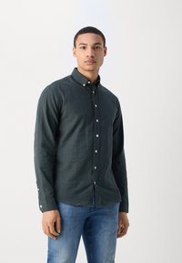 Dark green button-up shirt made of textured fabric, featuring a collar and silver buttons, worn with blue jeans.