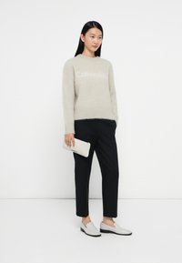Lichtgrijze fuzzy trui met "Calvin Klein" logo, gecombineerd met zwarte taps toelopende broek en witte instappers. Houdt een gestructureerde witte clutch vast.
