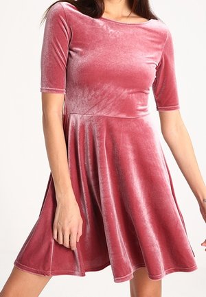 Kobieta ubrana w różową aksamitną sukienkę z krótkim rękawem, z dopasowaną talią i rozkloszowanym dołem, stojąca na jednolitym tle.