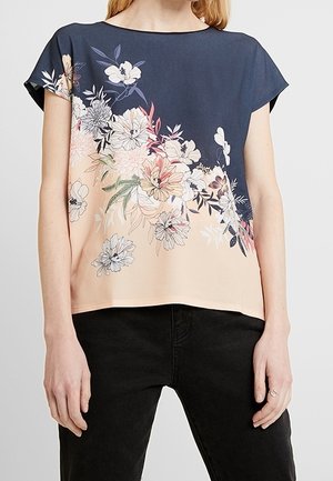 Vrouw draagt een top met korte mouwen met een marineblauwe bovenkant en een perzikkleurige onderkant met bloemenprint, gecombineerd met een zwarte broek.