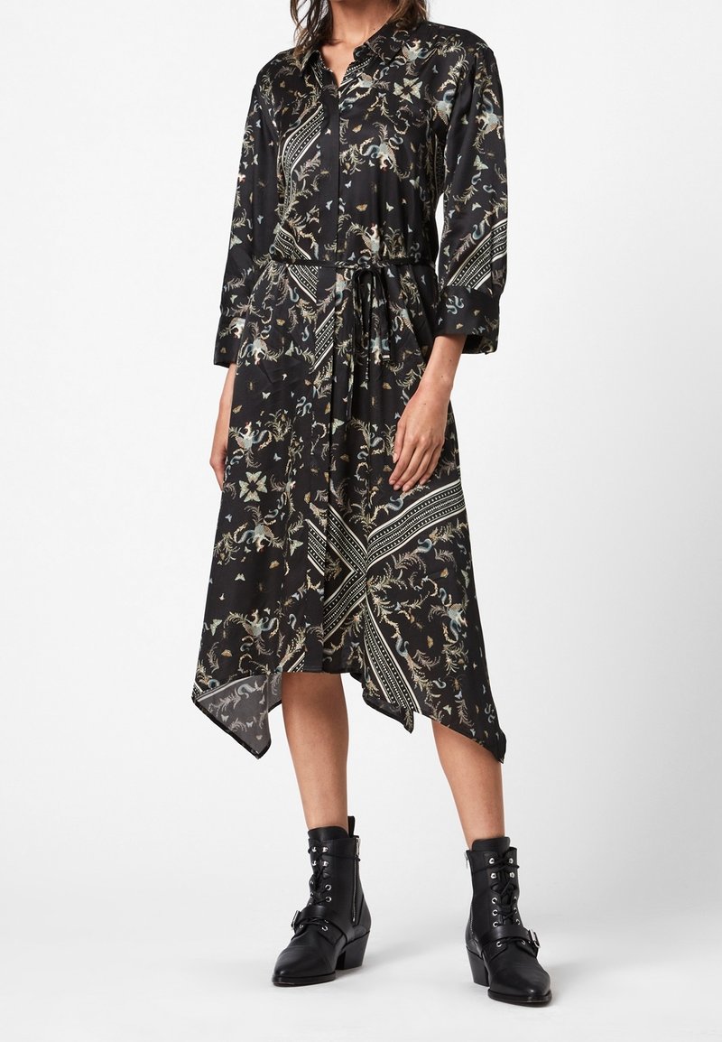 Femme portant une robe longue noire à motif floral, manches longues, avec une ceinture à nouer à la taille et des bottines noires à lacets.