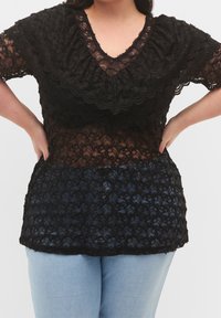 Svart spetsoberdel med vågig design, V-ringning och korta ärmar, lager över en genomskinlig väv med bred, texturerad nederkant.