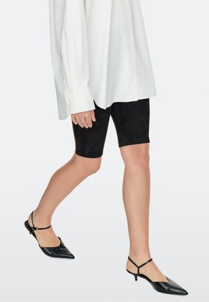 Personne portant un short noir ajusté mi-cuisse, une chemise blanche oversize à boutons et des escarpins noirs ouverts à l'arrière avec un talon kitten pointu, en train de marcher.