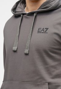 Sivá mikina s kapucňou a sťahovacou šnúrkou, s textúrovanými tkanými šnúrkami. Na hrudi čierne logo EA7 Emporio Armani.