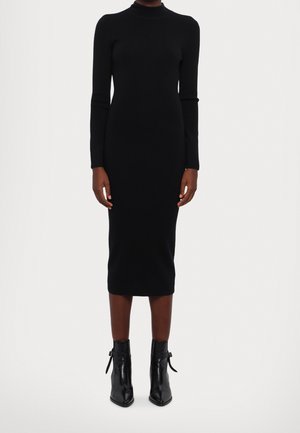 Žena oblečená v přiléhavých, černých midi šatech s dlouhým rukávem, doplněných černými špičatými kotníkovými botami na bílém pozadí.