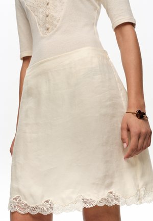 Mujer que lleva una falda de raso color crema con ribete de encaje en el dobladillo y una blusa a juego con mangas hasta el codo y detalles bordados.