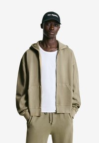 Khaki zip-up hoodie with a relaxed fit, paired with a white t-shirt and matching drawstring pants. Black cap with “try again.” text.