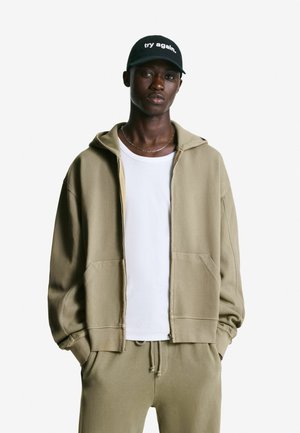 Khaki zip-up hoodie with a relaxed fit, paired with a white t-shirt and matching drawstring pants. Black cap with “try again.” text.