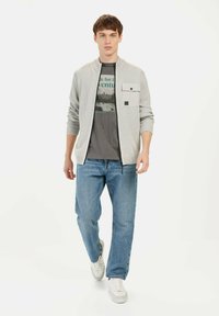 Lichtgrijze cardigan met geribde manchetten, ritssluiting en een borstzak, gedragen over een donkergrijze T-shirt met print. Gemaakt met lichtblauwe jeans en witte sneakers.