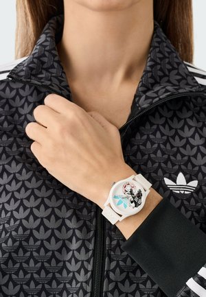 Ceas de mână alb cu un design grafic ce include o ilustrație detaliată și o carcasă rotundă, purtat pe o mână sub o jachetă neagră cu model.