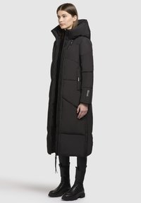 Manteau d'hiver noir long avec un col haut, fermeture éclair, texture matelassée et poches latérales. Associé à des bottes de combat noires.