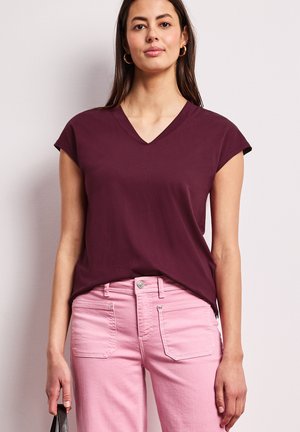 Frau trägt ein bordeauxrotes ärmelloses Top mit V-Ausschnitt und hellrosa Jeans, hält eine schwarze Handtasche vor neutralem Hintergrund.