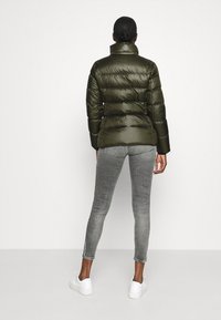 Vadderad olivgrön jacka med glänsande textur, hög krage och horisontell sömnadsdetalj. Matchas med gråa tajta jeans och vita sneakers.