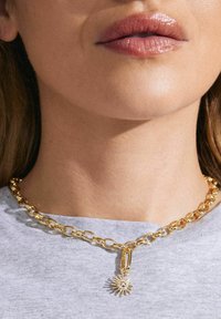 Auksinė grandinėlė su didelėmis grandinėmis ir saulės spindulio pakabuku su skaidriu akmeniu, nešiojama prieš pilką marškinį.