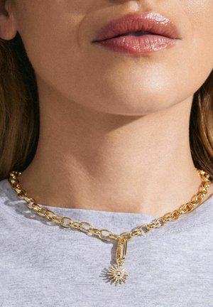 Kuldne kett, millel on suured lingid ja päikesepaiste pendant, millel on selge gemstone detail, kantud halli särgi kohal.