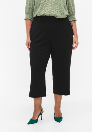 Black cropped trousers with a smooth texture, high waist, and side pockets, paired with green stiletto heels and a patterned green blouse.