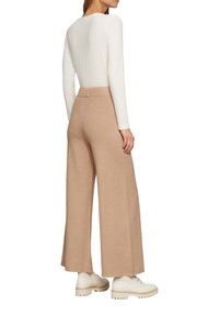 Beige Weitbein-Hosen mit glatter Textur; kombiniert mit einem weißen Langarmoberteil und cremefarbenen Schuhen mit dickem Profil.