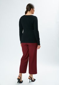 Schwarzer langärmeliger Pullover mit einer figurbetonten Passform, kombiniert mit burgunderfarbenen, weiten Hosen. Sichtbare schwarze High-Heels-Sandalen. Neutraler Hintergrund.