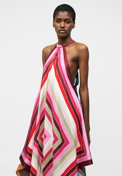 Γυναίκα που φοράει φόρεμα με λαιμόκοψη halter και τολμηρές διαγώνιες ρίγες σε ροζ, κόκκινο, άσπρο, πράσινο και βουργουνδί χρώματα σε απλό φόντο.