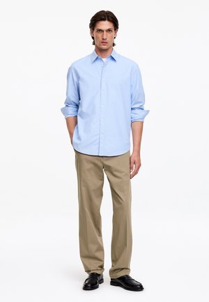 Jeune homme debout portant une chemise bleu clair boutonnée, un pantalon beige et des chaussures noires sur un fond blanc uni.