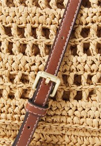Primo piano di una borsa di paglia intrecciata con una tracolla in pelle marrone dotata di una fibbia dorata e cuciture bianche a contrasto, che mette in evidenza il materiale testurizzato.