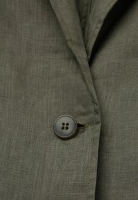 Kaki linneväv med en slät yta, som har en rund knapp med fyra hål och en liten trådslinga för att fästa.